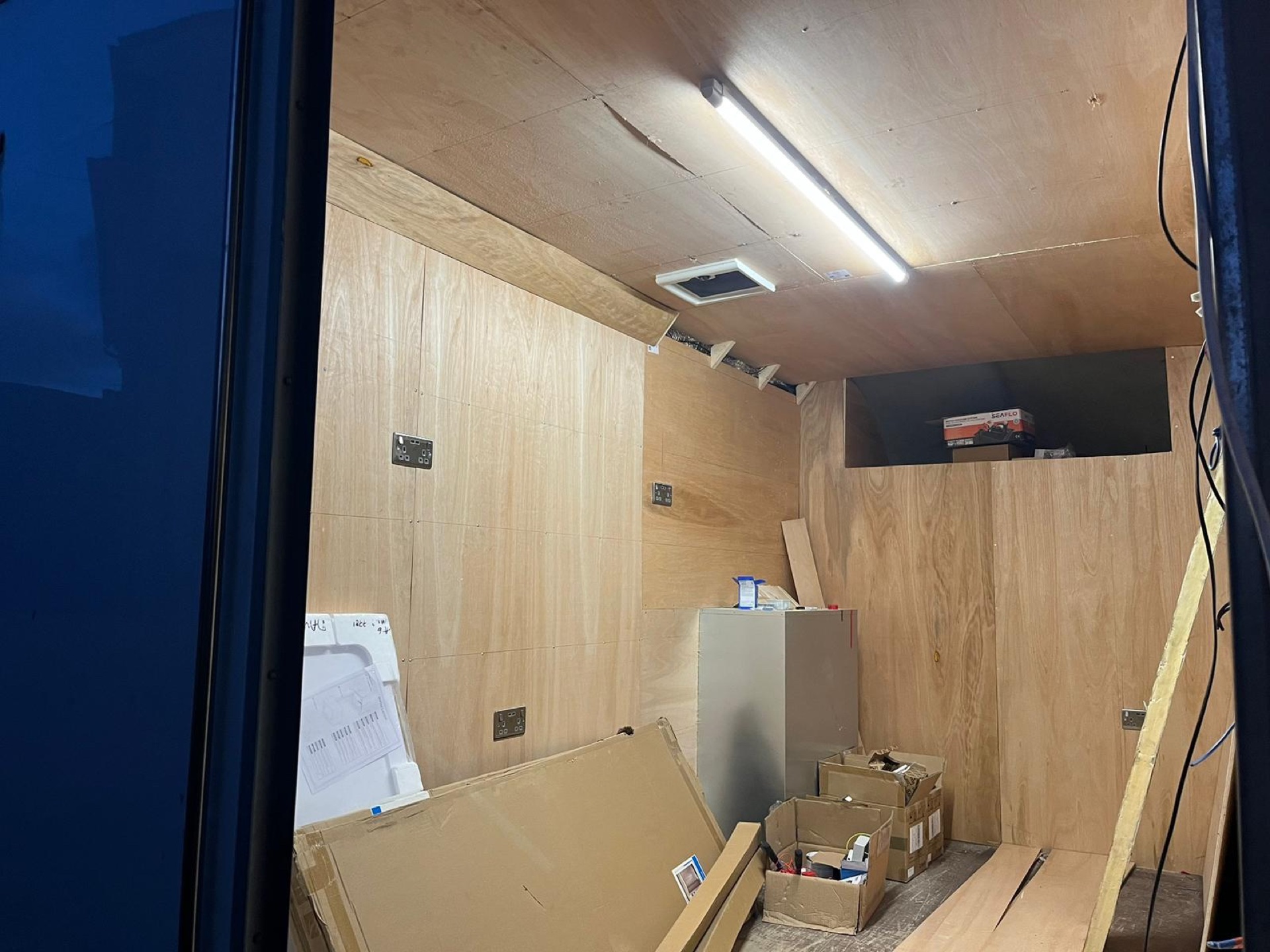 Van interior lined with plywood, strip light on, boxes of fittings and plans taped to the wall