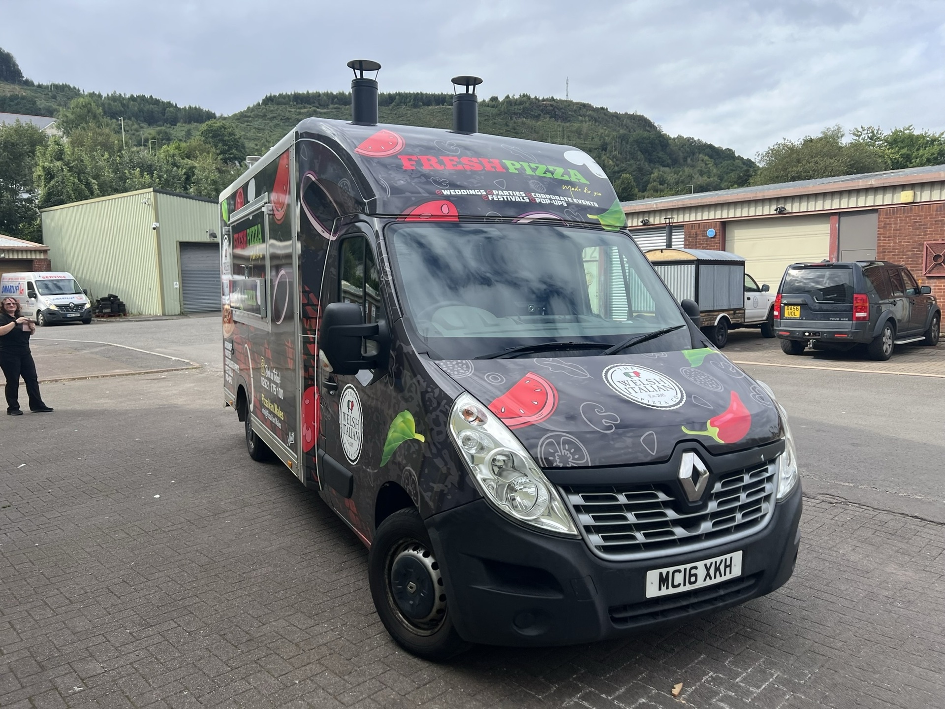 The finished pizza van wrapped in charcoal with chilli and tomato artwork, hillside behind