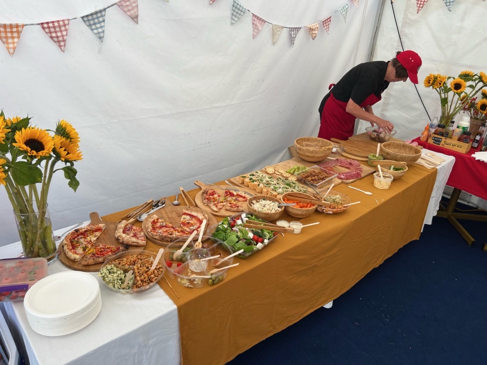 A long grazing table dressed with bunting and sunflowers, boards of pizza being laid out