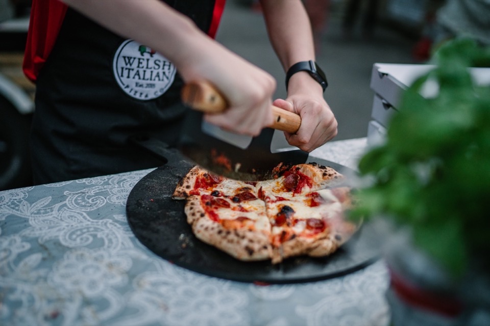A freshly fired pepperoni pizza being sliced on a black board at an event