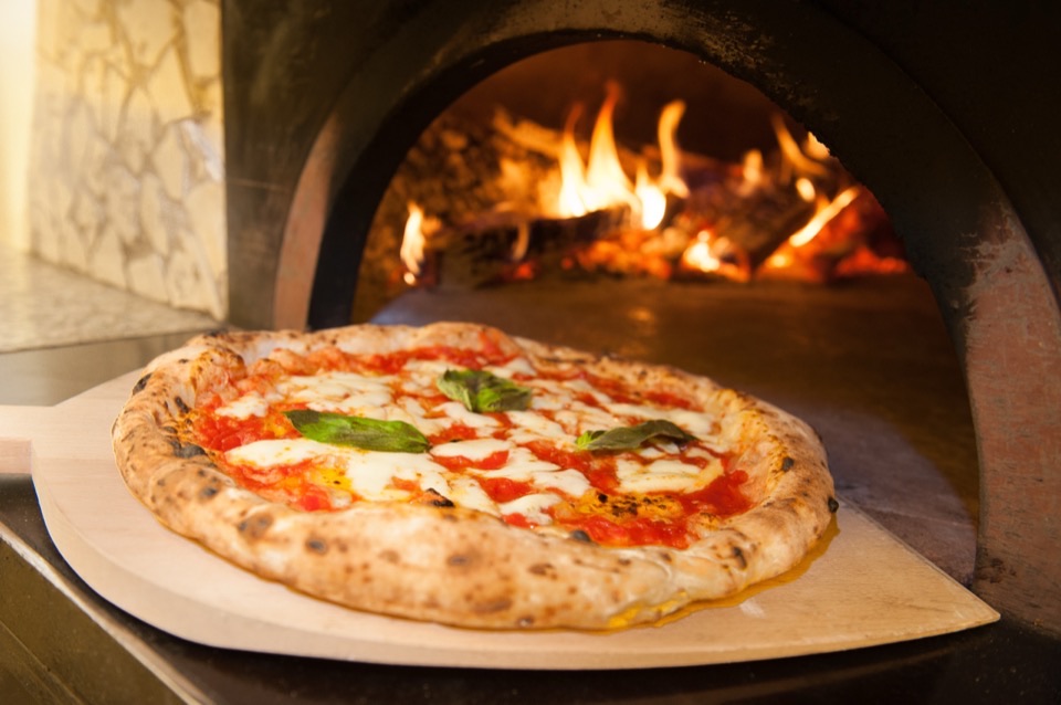 A margherita pizza on a wooden peel beside a lit pizza oven