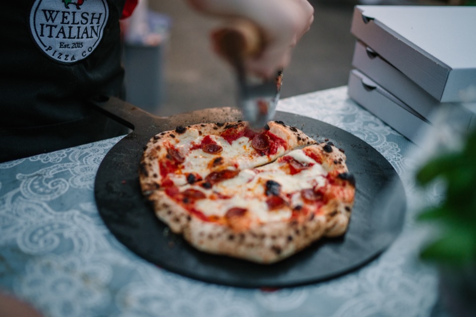 A pepperoni pizza being sliced beside stacked boxes, the crew apron showing the Welsh Italian Pizza Co stamp, at Alana and David's wedding